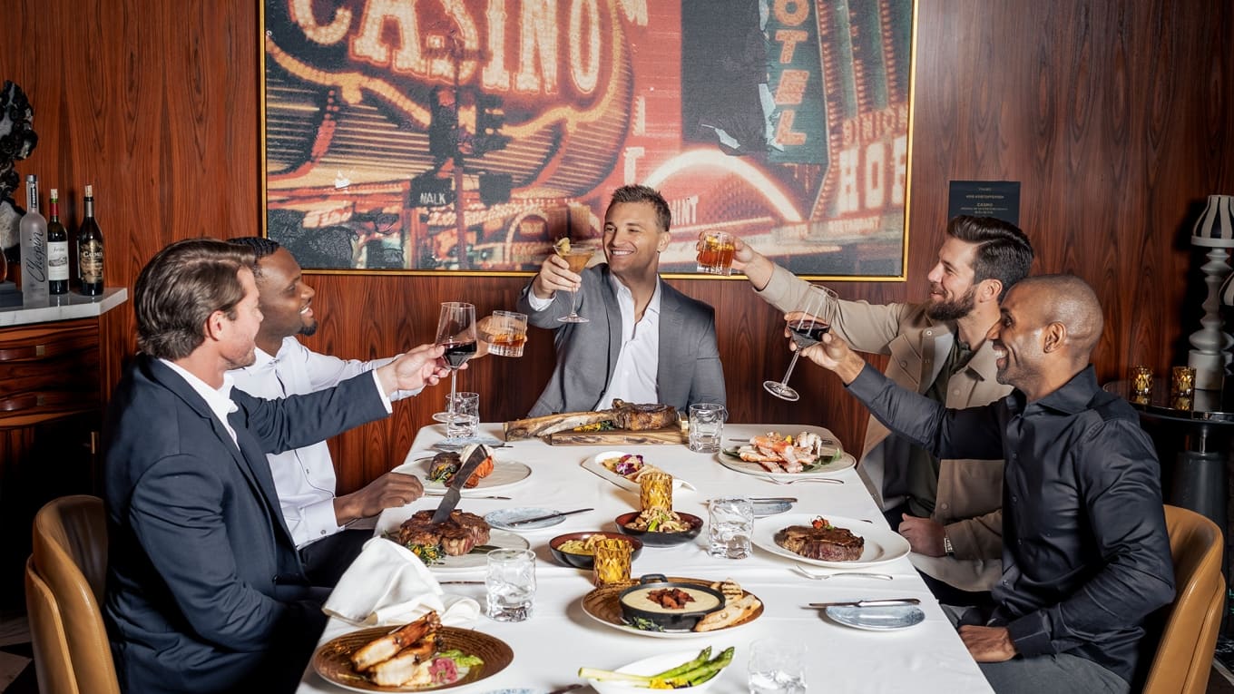 Five men raising a toast over steaks and shellfish at a long table