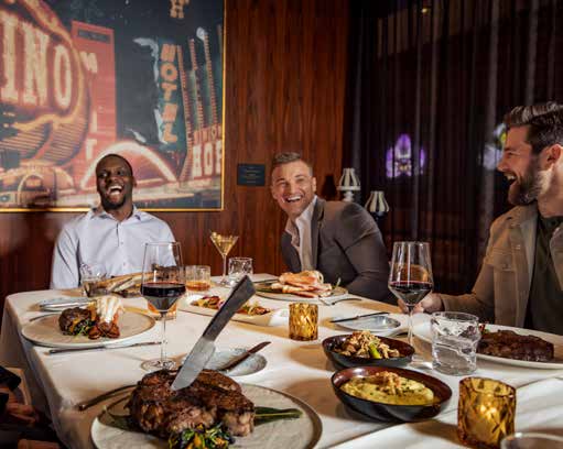 Three men laughing over steaks and red wine at a white linen table