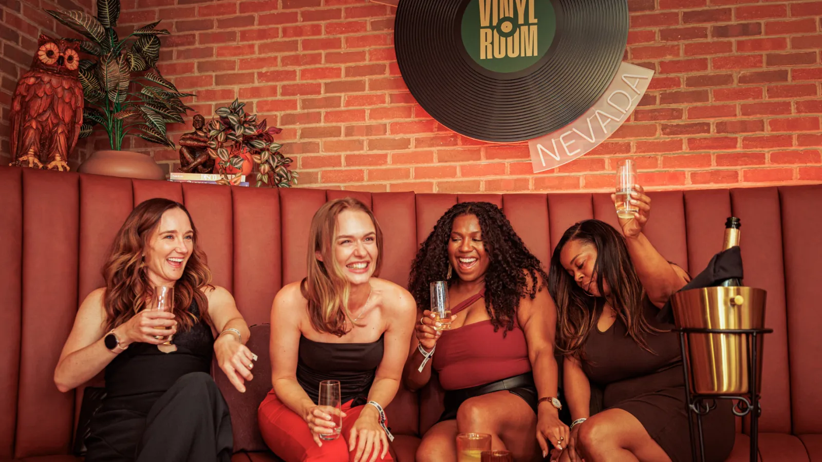 Four friends laughing on a curved leather banquette at Vinyl Room Las Vegas beneath a large Vinyl Room record and a Nevada sign