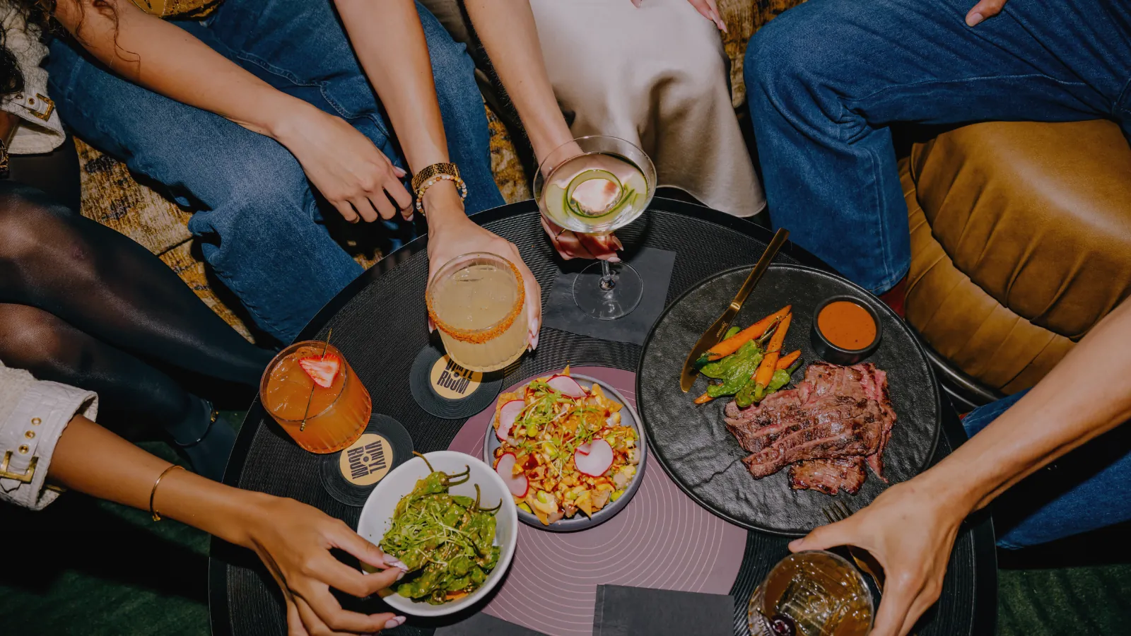 Overhead view of a low table at Vinyl Room set with sliced steak, poke nachos, shishito peppers and four cocktails