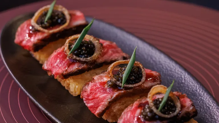 Slices of rare wagyu beef on crispy rice topped with caviar and scallion on a dark plate